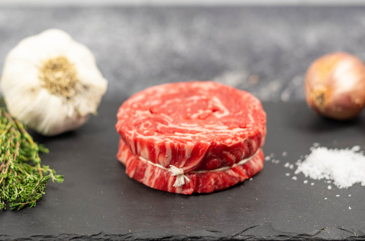 Prime Ribeye Cap - Rolled 10 oz, showcasing marbled meat on a slate background, accompanied by garlic, shallots, and herbs.