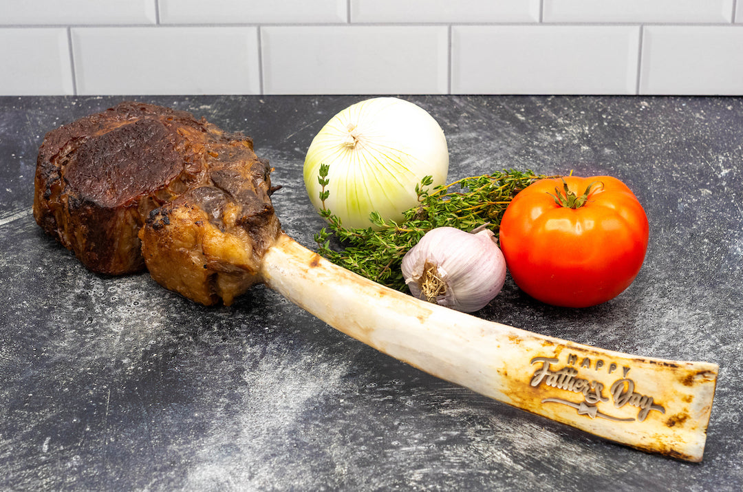 Engraved "Happy Father's Day" dry-aged USDA Prime Black Angus tomahawk steak beside fresh vegetables, including onion, tomato, garlic, and herbs.